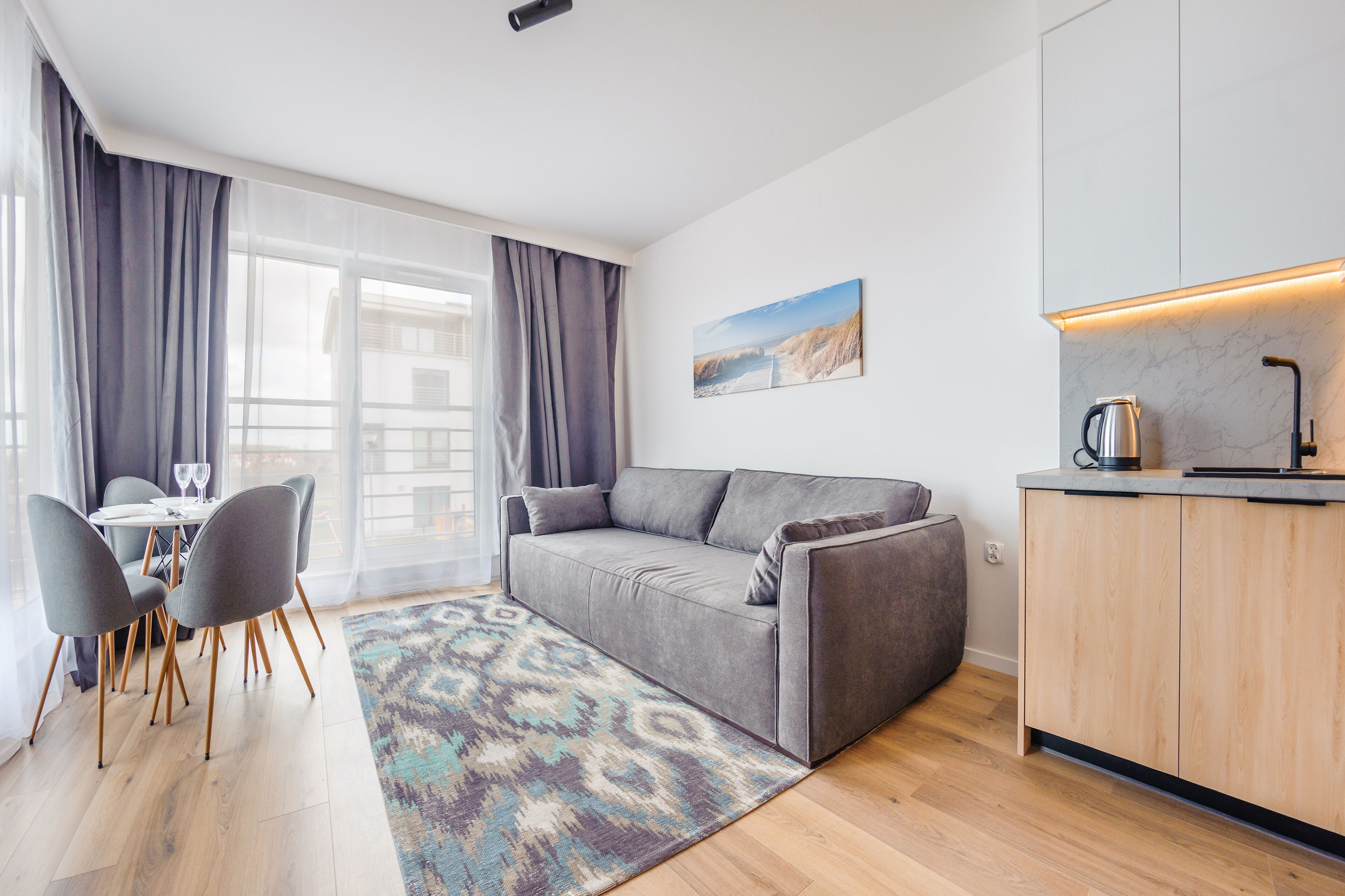 A modern living room with a grey sofa, a dining table with chairs, and a kitchen area with a coffee machine.