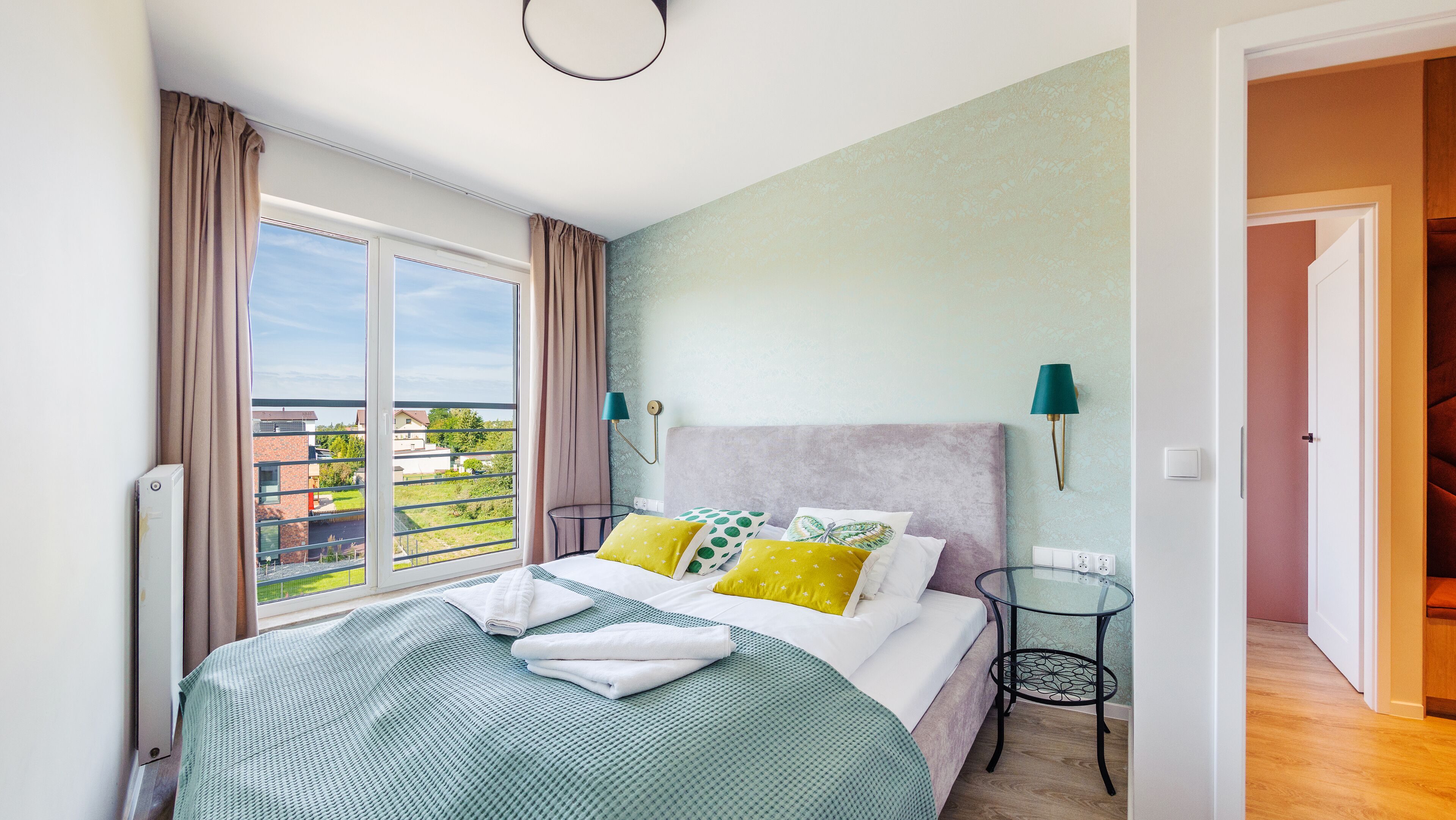 A bedroom with a large bed, a view of the city, and a small table with a lamp.