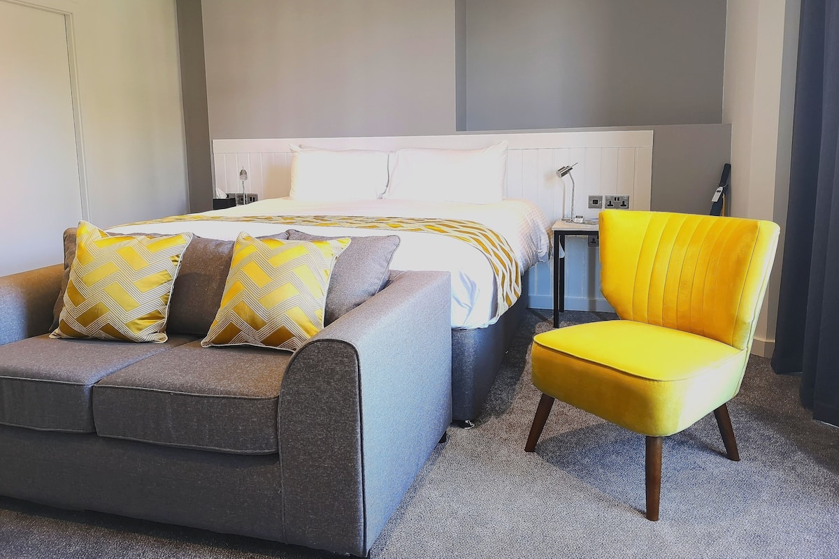 A hotel room with a bed, a sofa, a chair, and a desk with a lamp.