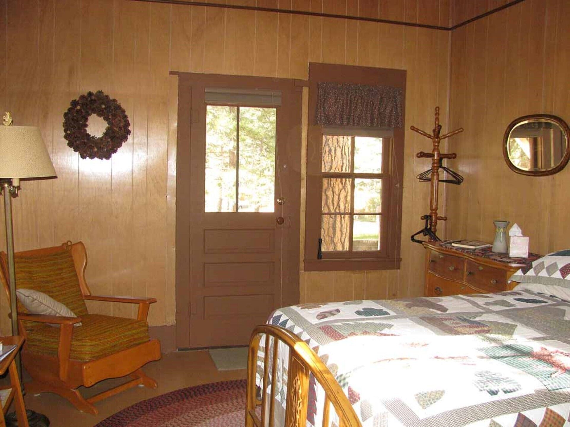 Habitación con paredes y puerta de madera, ventana con vistas, cama con colcha, una silla, tocador, lámpara y espejo.