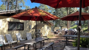 A patio area with outdoor tables and chairs, covered by red umbrellas, and surrounded by trees.