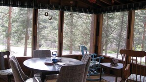 A cozy cabin interior with a round table and chairs, large windows offering a forest view, and a rustic ceiling with hanging decorations.
