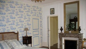 Un dormitorio con cama, mesita de noche, lámpara, chimenea, espejo y puerta que da a otra habitación.