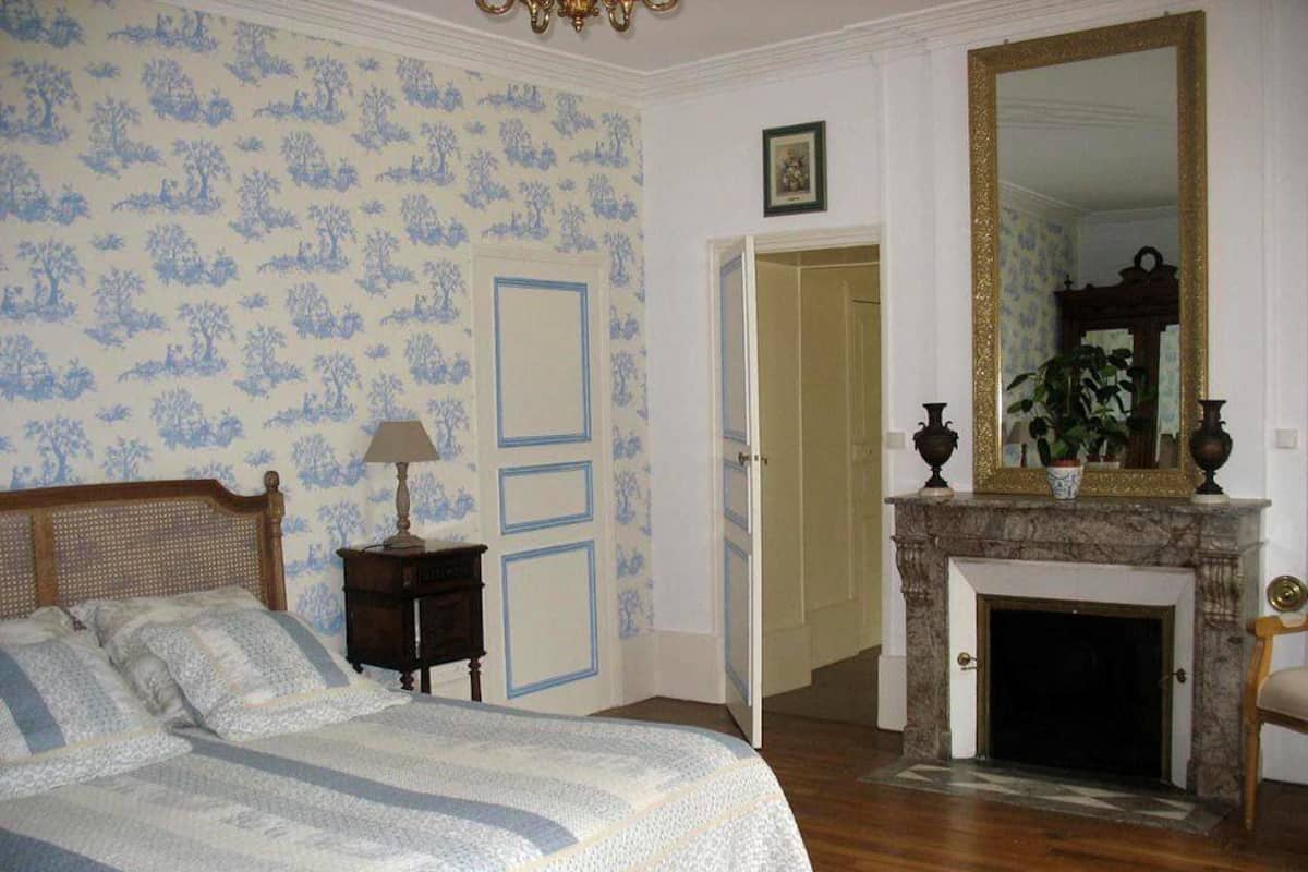 Une chambre à coucher comprenant un lit, une table de chevet, une lampe, une cheminée, un miroir et une porte donnant sur une autre pièce.