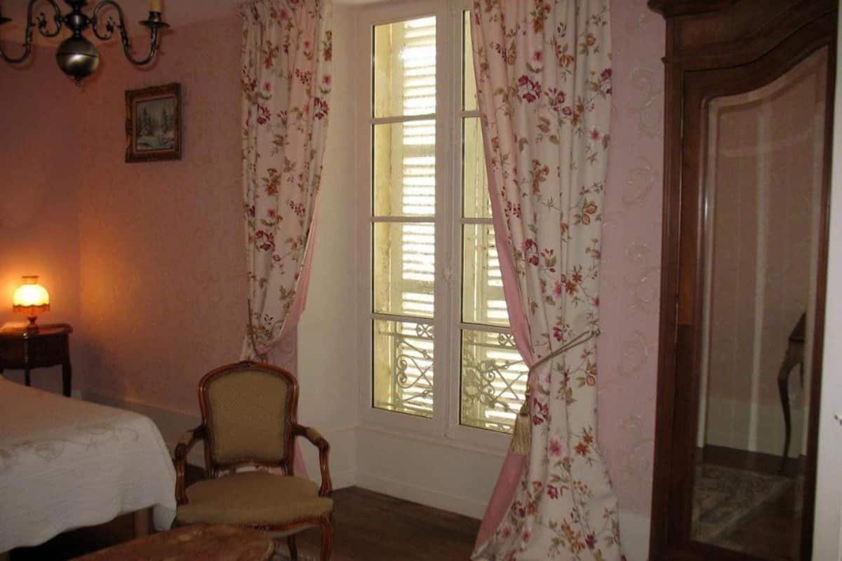 Une pièce comprenant un lit, une chaise, une table et une fenêtre ornée de rideaux à motifs floraux.