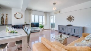 Una sala moderna con sofá, mesa de centro y televisión. Hay una zona de comedor con sillas y mesa. La habitación tiene ventanas grandes y una lámpara de techo.