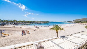 Una playa con cielo azul despejado, orilla de arena y algunas personas. Se ven sombrillas y sillas de descanso.
