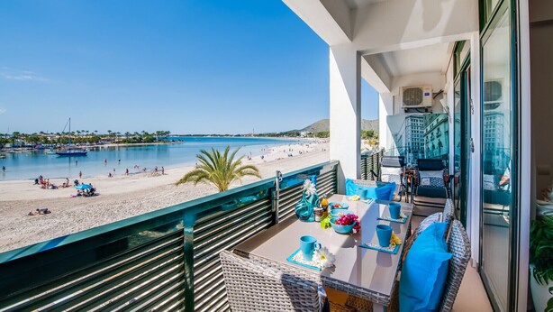 Un balcón con vistas a una playa y al mar, amueblado con una mesa y sillas, y un ventanal de vidrio que da a un paisaje urbano.