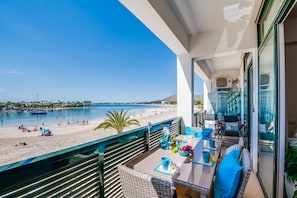 Ein Balkon mit Blick auf einen Strand und das Meer, ausgestattet mit einem Tisch und Stühlen sowie einer Glastür, die auf eine Stadtansicht führt.
