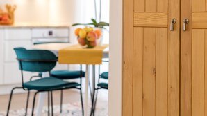 Una puerta de madera con dos pomos plateados, un comedor con una mesa amarilla y sillas verdes, y una cocina con armarios blancos y una lámpara colgante.