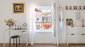 Una habitación con un ventanal grande, un escritorio con un florero, un taburete pequeño y un mueble blanco con objetos decorativos.