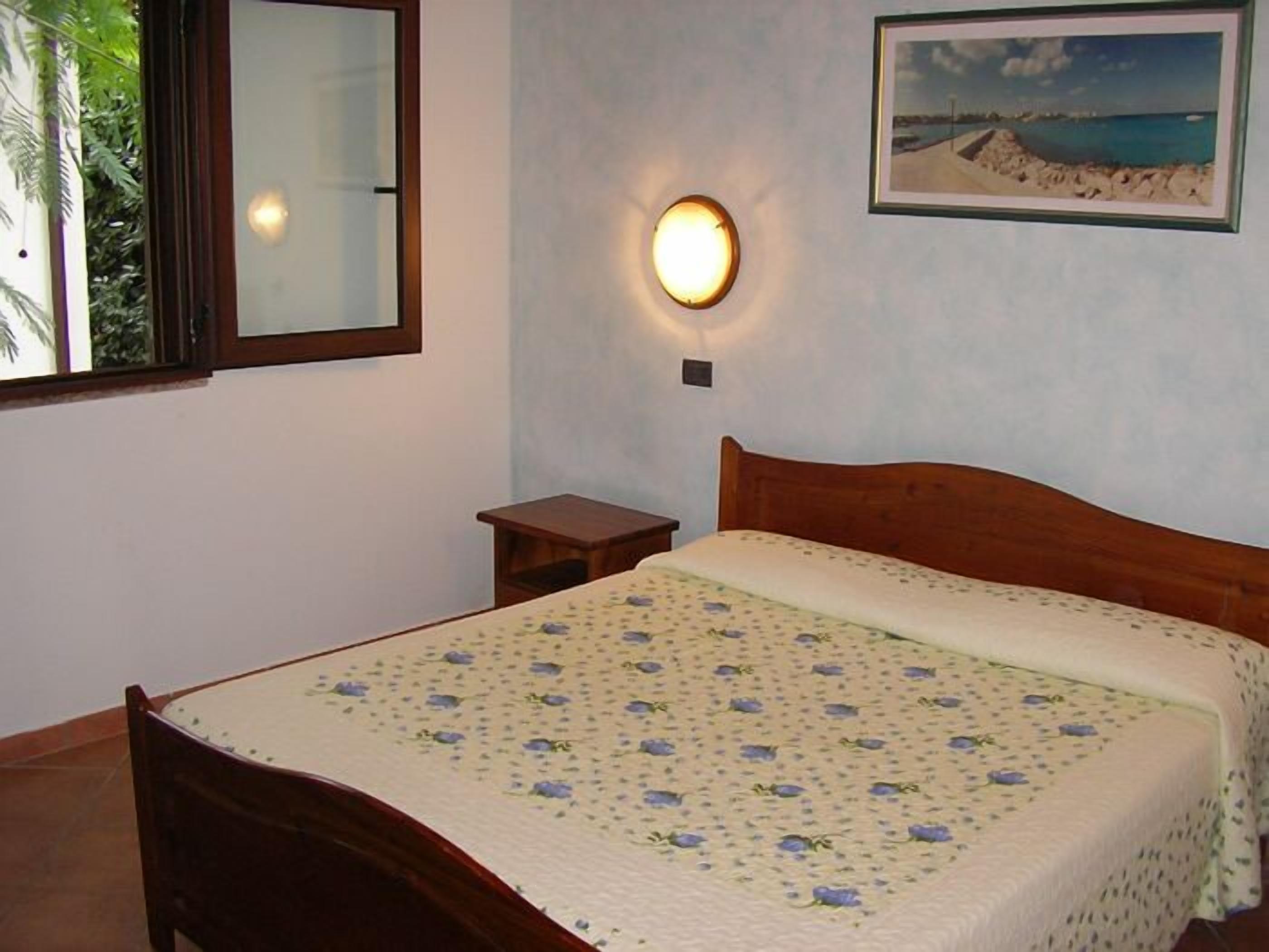 A bedroom with a wooden bed, a nightstand, a wall-mounted lamp, a window with a view of greenery, and a framed picture on the wall.