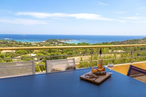 Una mesa con una botella, un vaso y comida en un balcón con vistas al paisaje costero.