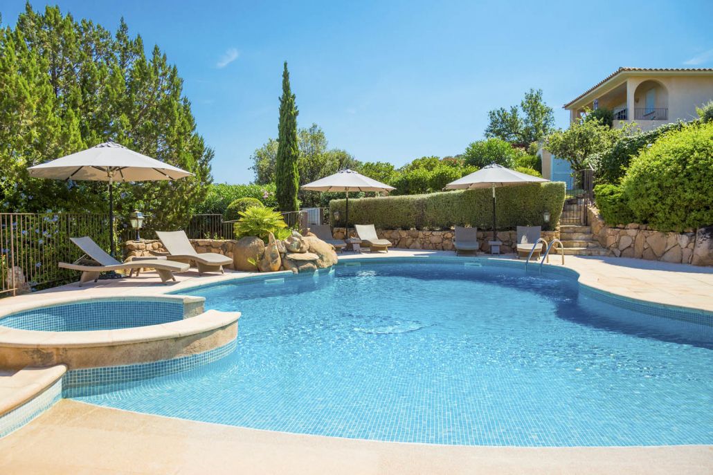 Ein Swimmingpool mit Liegestühlen und Sonnenschirmen, umgeben von Grünflächen und einer Steinmauer.