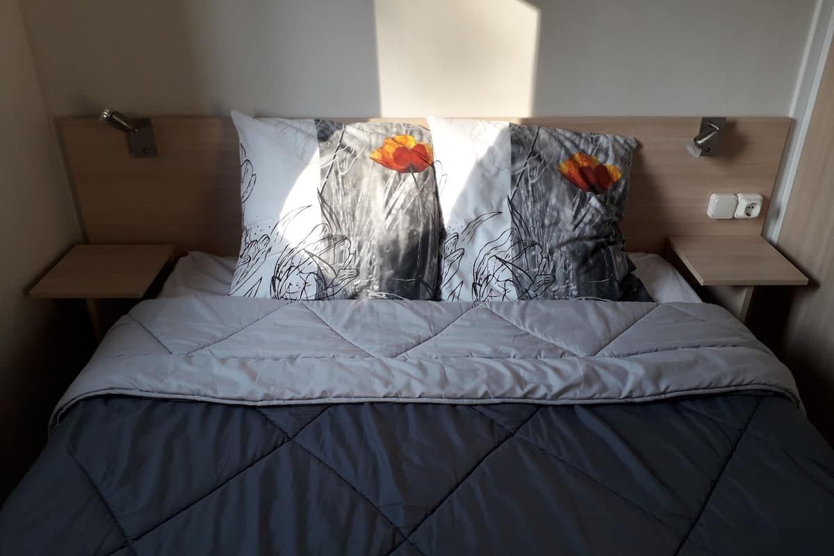 Un lit bien fait, avec une couette grise matelassée, deux oreillers à motifs floraux et une tête de lit en bois.