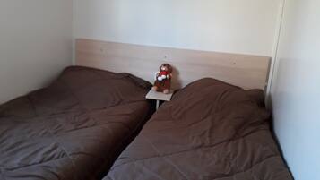 Une chambre avec deux lits, chacun recouvert d’une couette marron. Il y a une étagère au-dessus des lits et une petite table sur laquelle est posé un ours en peluche.