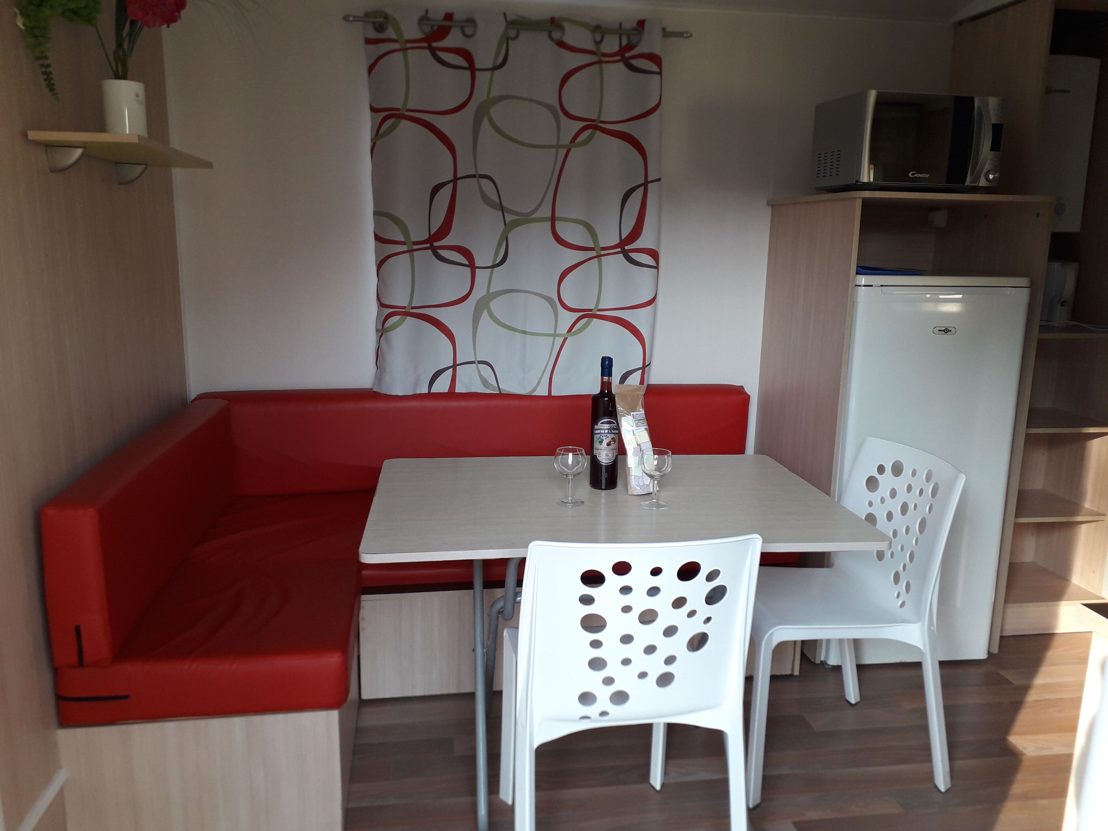 Un espace de vie compact avec un canapé rouge, une table blanche et deux chaises blanches. Une bouteille et des verres sont posés sur la table. On peut apercevoir un four à micro-ondes et un réfrigérateur à l’arrière-plan.