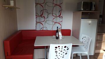 Un espace de vie compact avec un canapé rouge, une table blanche et deux chaises blanches. Une bouteille et des verres sont posés sur la table. On peut apercevoir un four à micro-ondes et un réfrigérateur à l’arrière-plan.