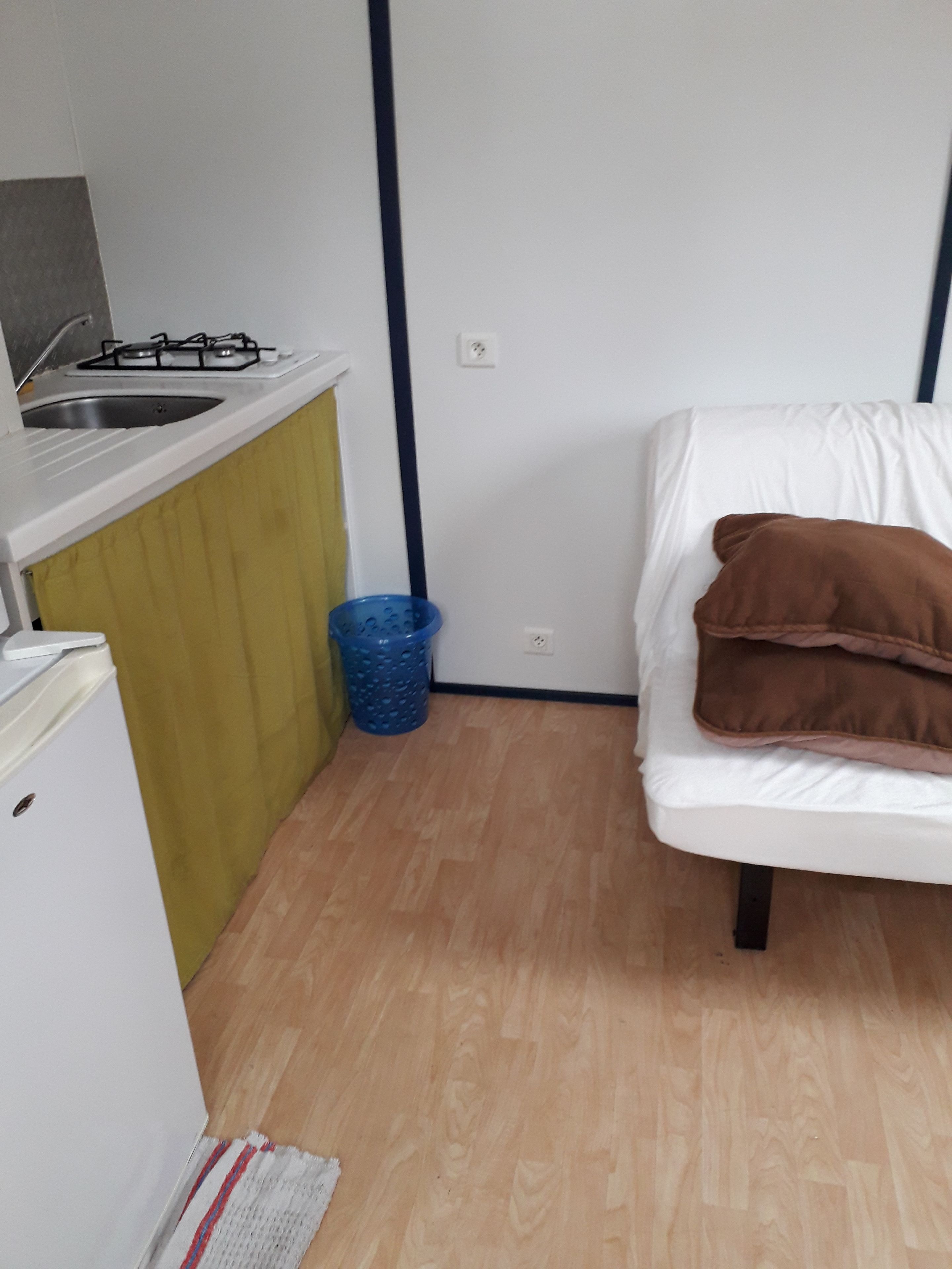 A compact room with a kitchenette on one side, a bed with pillows, and a blue trash bin.
