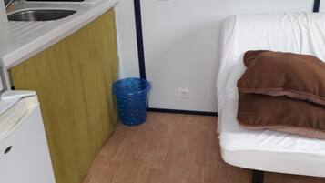 A compact room with a kitchenette on one side, a bed with pillows, and a blue trash bin.