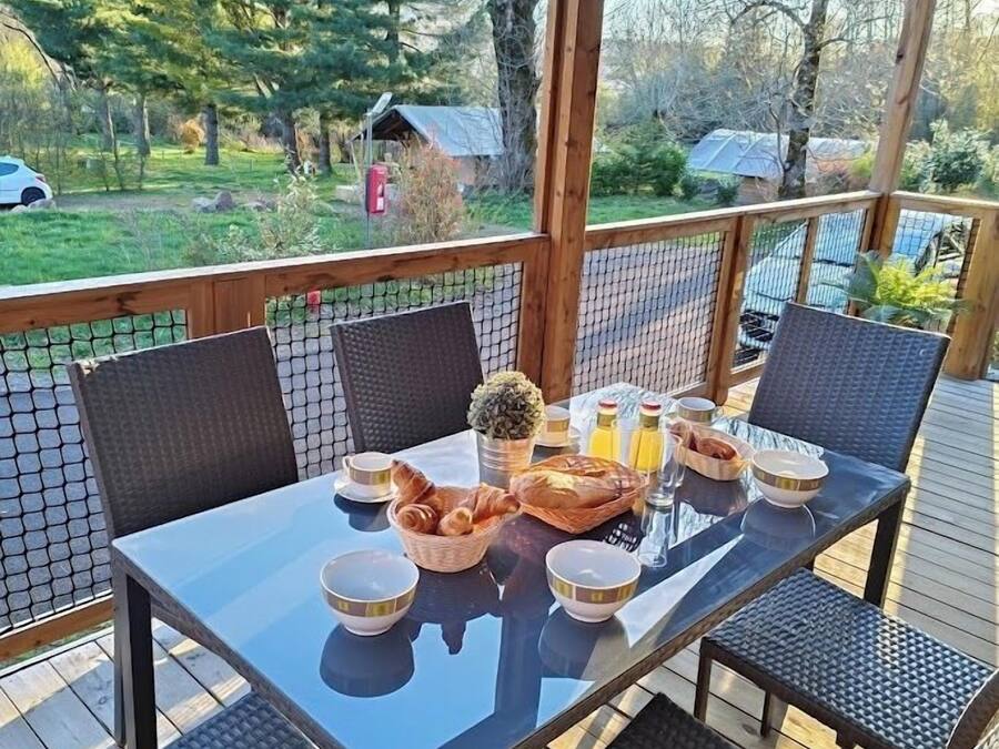A covered outdoor dining area with a table set for breakfast, surrounded by wicker chairs, overlooking a garden and a road.