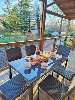 A covered outdoor dining area with a table set for breakfast, surrounded by wicker chairs, overlooking a garden and a road. - Camping la Marmotte (La Bourboule)