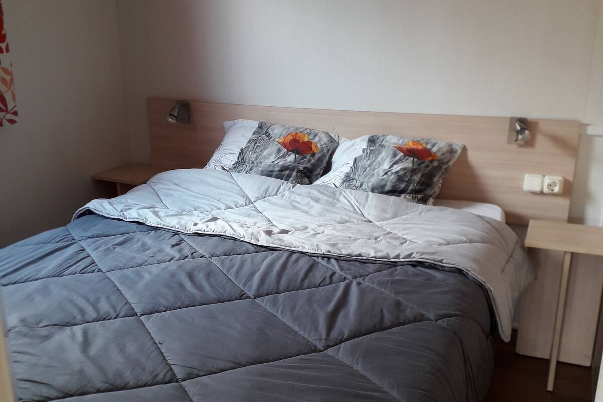Un lit bien fait, avec une couverture grise, une tête de lit en bois et une table de chevet surmontée d’une lampe.