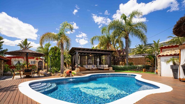 A swimming pool with a wooden deck, a covered seating area, and palm trees.