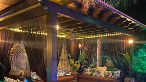 A covered outdoor seating area with a Buddha statue, cushioned seating, and potted plants.