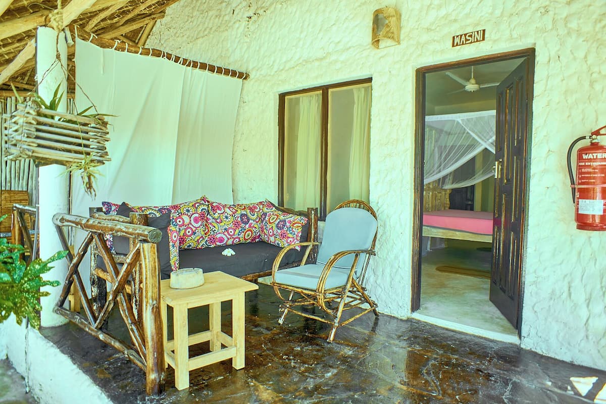 Una habitación rústica con una silla de mimbre, una mesa de madera y un sofá con una manta colorida. Se ve una puerta con la leyenda "WASINI", y un extintor está colgado en la pared.