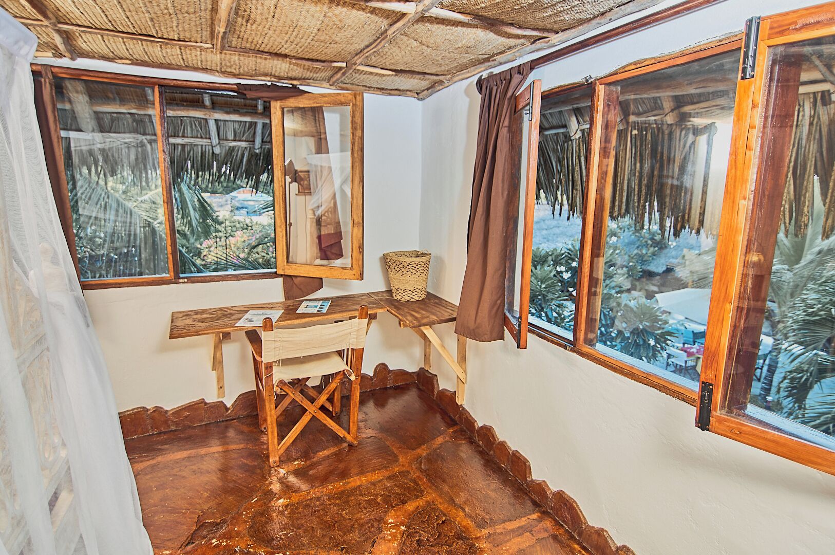 A rustic room with a wooden desk, a mirror, and a view of a tropical landscape through large windows.