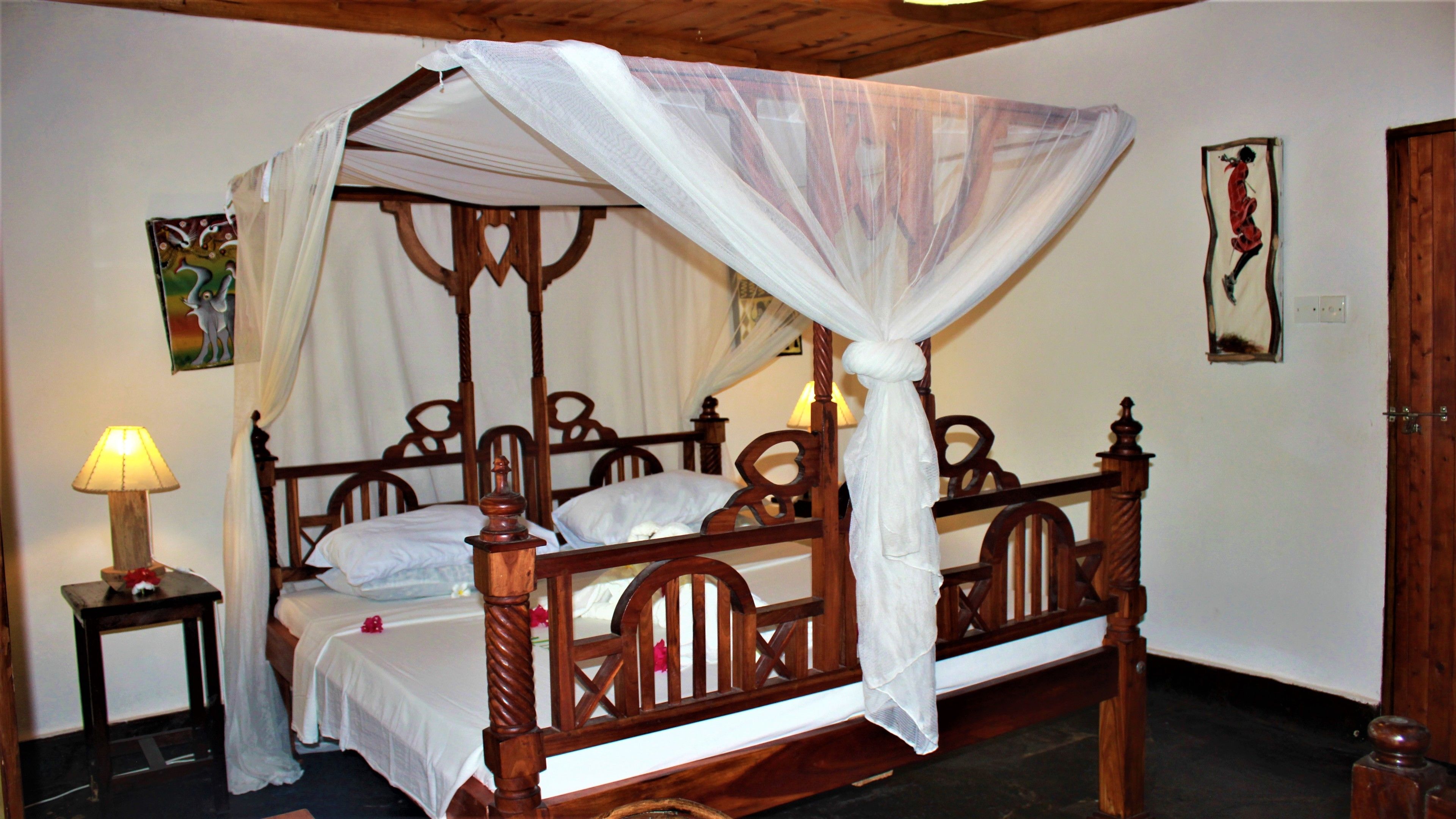 A four-poster bed with a canopy, wooden headboard, and white bedding. There is a bedside table with a lamp and a small decorative item.