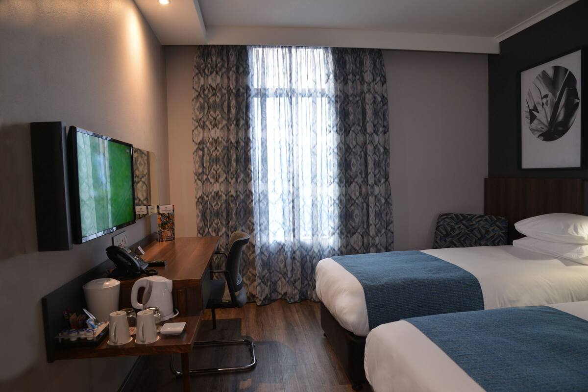 Een hotelkamer met twee bedden, een bureau met telefoon en koffiezetapparaat, een aan de muur bevestigde televisie en een raam met patroon gordijnen.