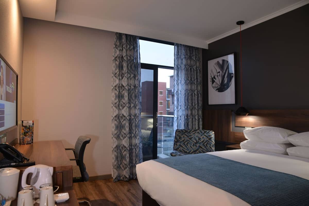 Een hotelkamer met een bed, een bureau met telefoon, een stoel, een koffiezetapparaat en uitzicht op de stad door grote ramen.