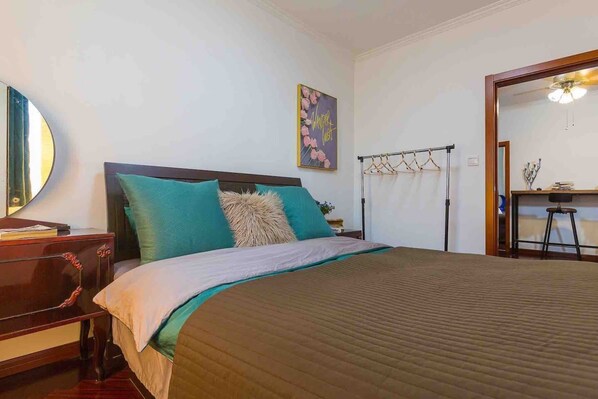 A neatly made bed with a brown quilt, teal and beige pillows, and a wooden headboard. A mirror hangs on the wall to the left, and a small table with a lamp is visible in the corner. - Splendiferous Apartment - Wang Jing (Beijing)