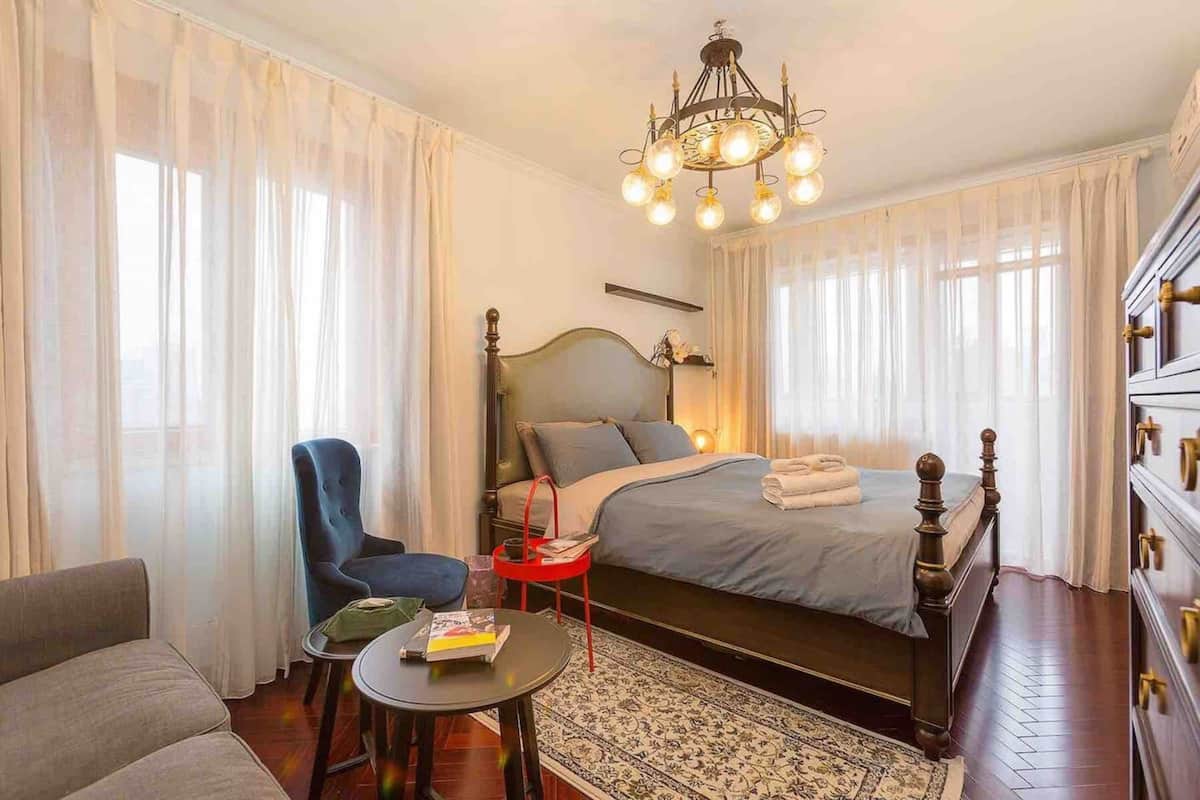 A hotel room with a bed, a chair, a small table, a chandelier, and a dresser.