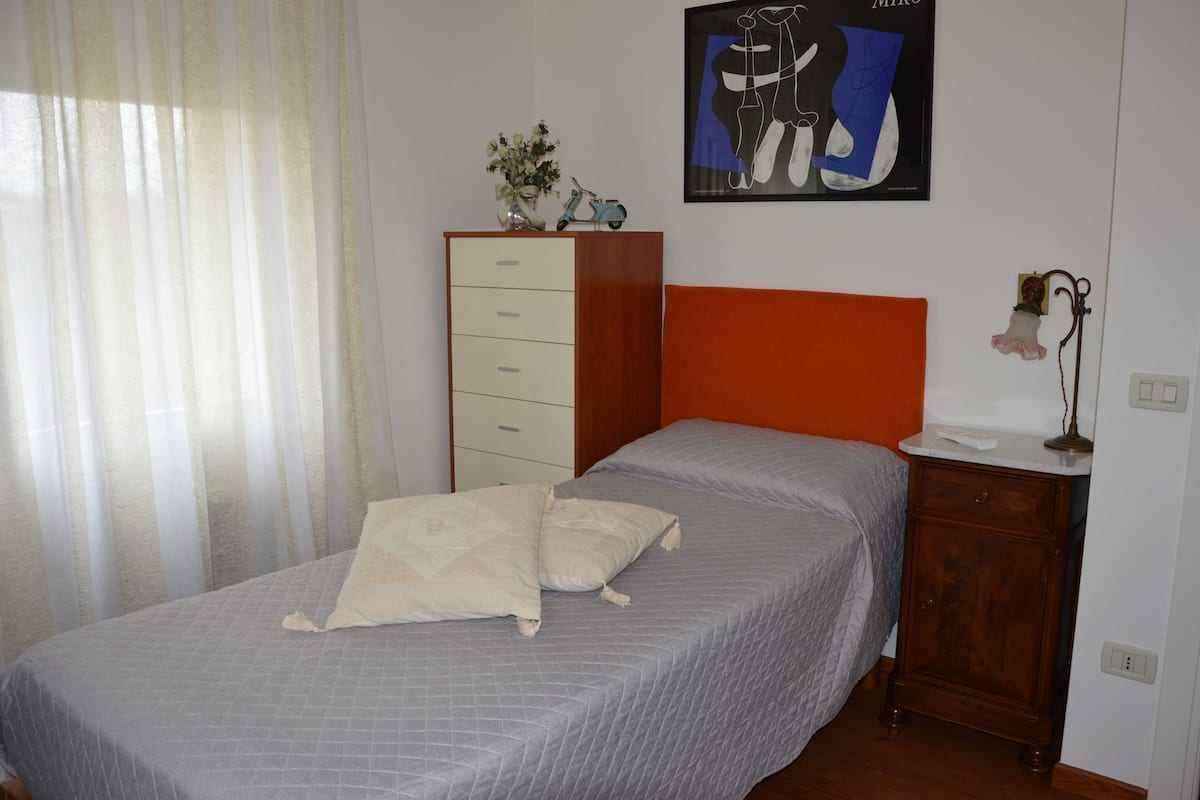 Une chambre à coucher comprenant un lit, une commode, une table de chevet, une œuvre d’art encadrée et une fenêtre avec des rideaux légers.