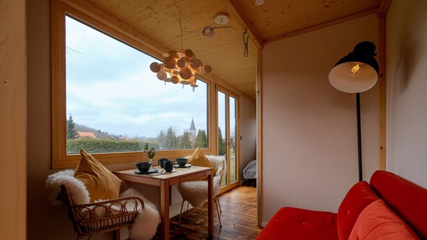 A cozy room with a wooden ceiling, a dining table with chairs, a red sofa, and a large window offering a view of the countryside. - Harzer Hof (Herzberg am Harz)