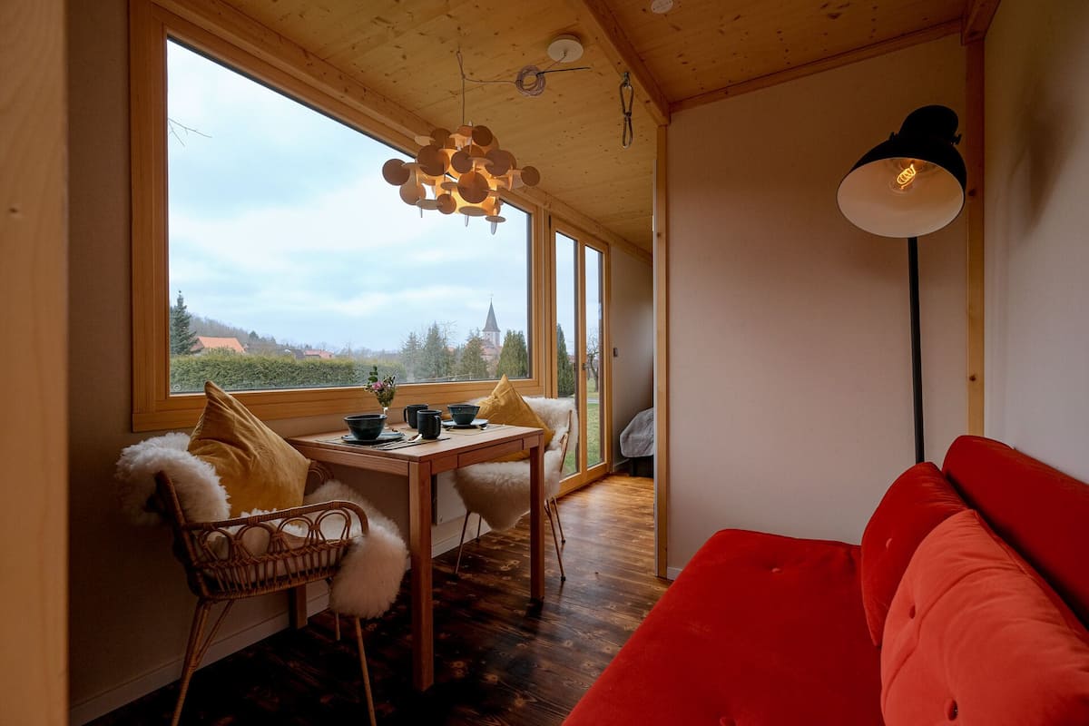 Une pièce chaleureuse avec un plafond en bois, une table à manger avec des chaises, un canapé rouge et une grande fenêtre donnant sur la campagne.