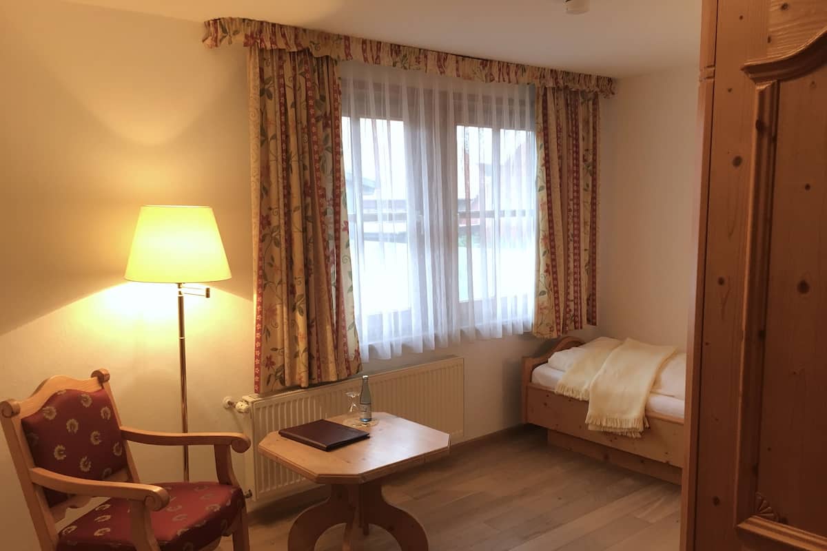 Une pièce confortable avec une chaise en bois, une petite table avec une lampe, un lit et une fenêtre avec des rideaux.