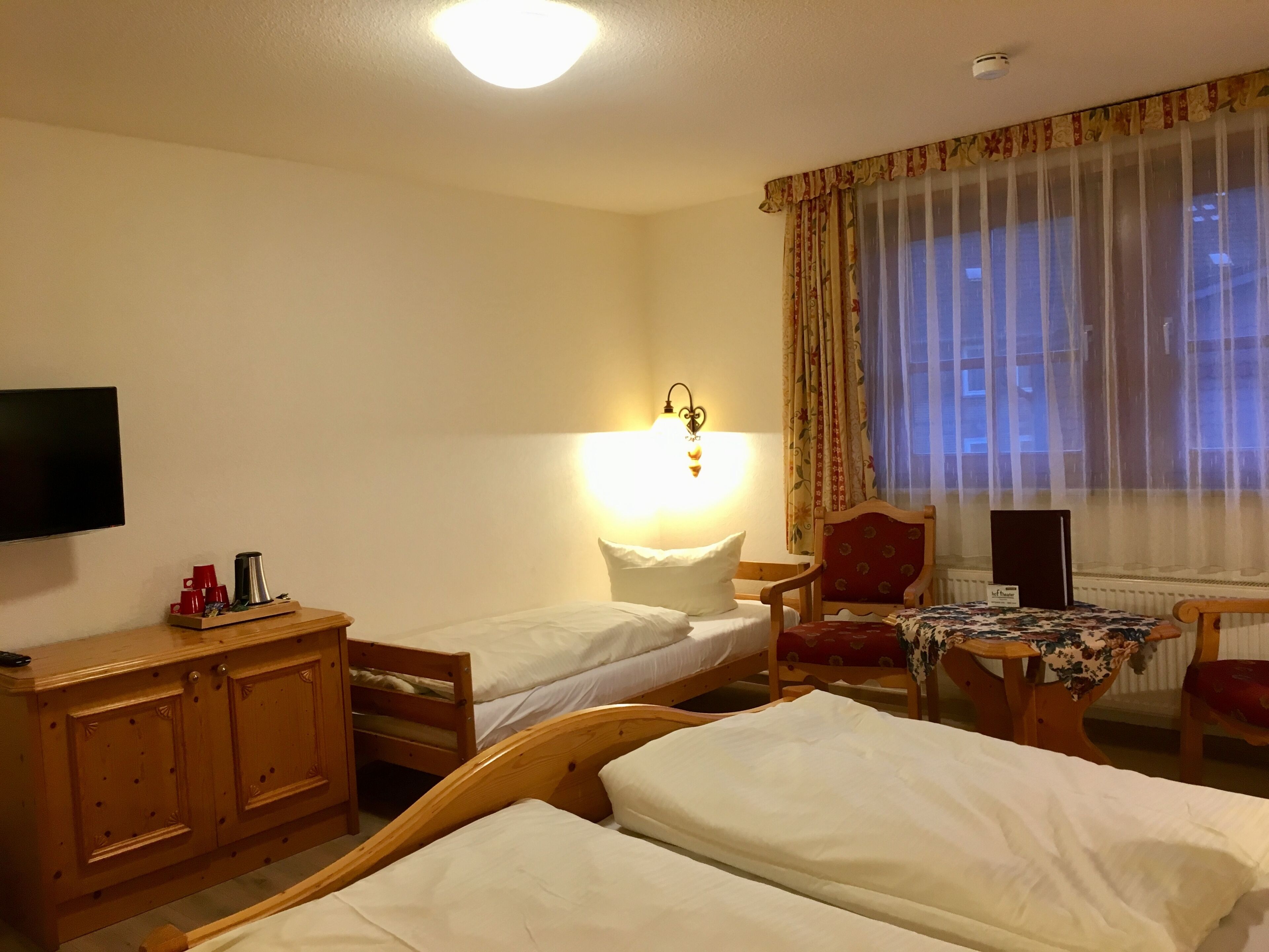 Ein Hotelzimmer mit zwei Einzelbetten, einem Holzkapfen, einem Schreibtisch mit Stuhl, einem Fernseher und einem Fenster mit durchsichtigen Vorhängen.