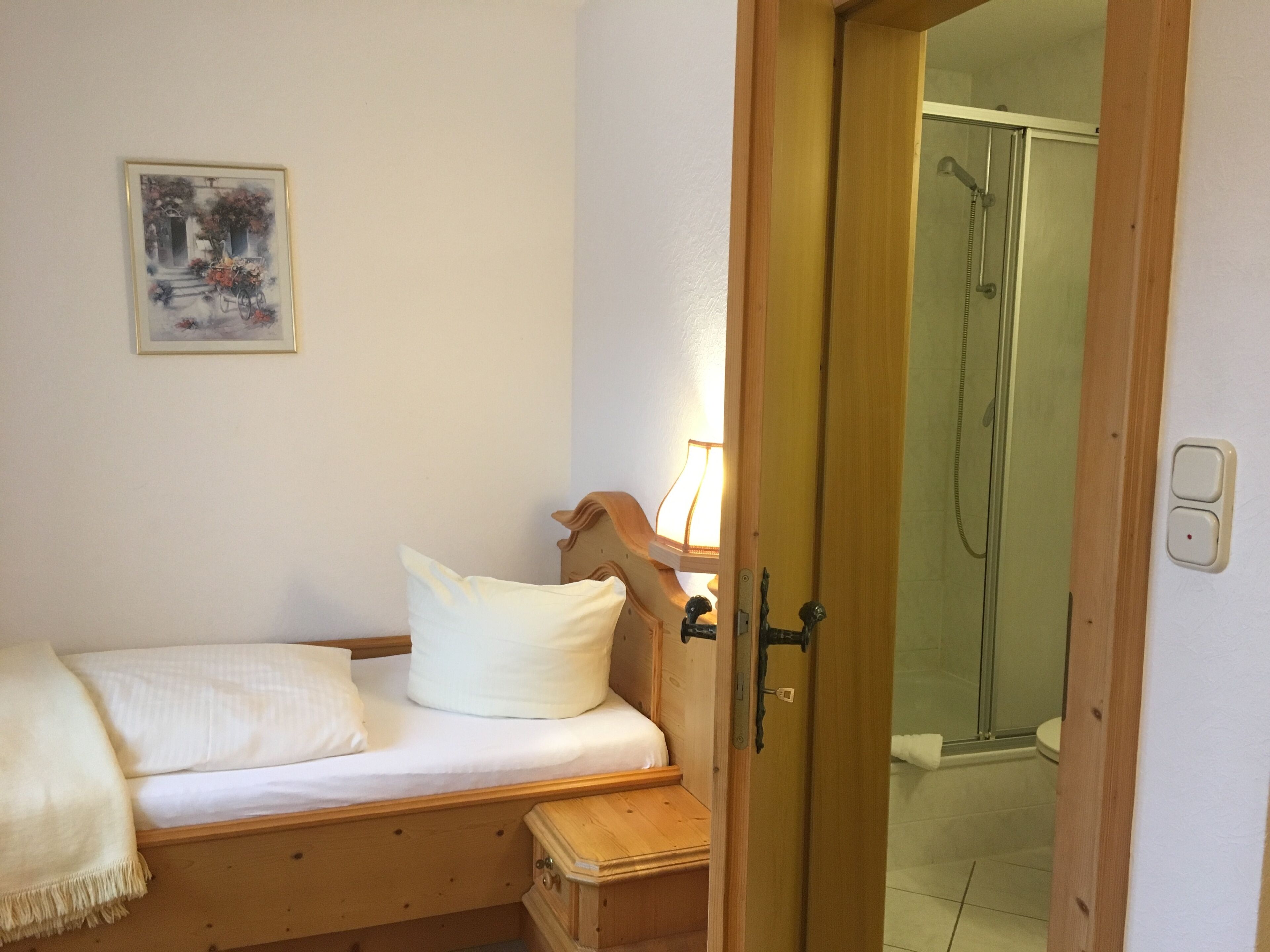A bedroom with a wooden bed, a bedside table, a lamp, and a shower area visible through an open door.