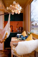 Un rincón comedor acogedor con una silla de mimbre, una mesa con cuencos azules y un cojín amarillo, y una lámpara colgante.