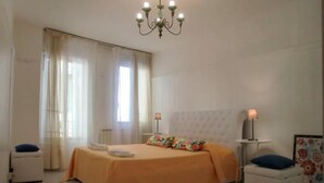 A bedroom with a large bed, a chandelier, a bedside table with a lamp, and a window with curtains. - Grimaldi Apartments - Jolie (Venice)