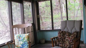 Une pièce avec deux chaises en bois, dont une avec un coussin à motifs floraux et l’autre avec un coussin à motifs camouflage, de grandes fenêtres donnant sur des arbres et un mur bleu.