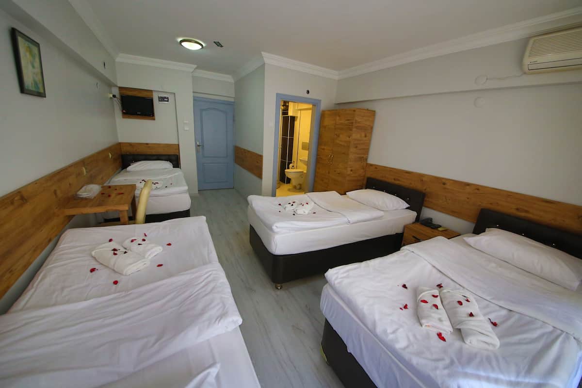 A hotel room with three beds, each with white linens and towels, and a wooden headboard. There is a wooden nightstand with a phone and a small table with a lamp. The room has a wooden floor and a door leading to a bathroom.