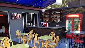 An outdoor seating area with wicker chairs and round tables, a bar with a bartender, and a refrigerator stocked with beverages. - Amelia Hotel Karen (Nairobi)