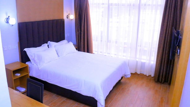 A hotel room with a large bed, a wooden nightstand, a TV, and a window with curtains. - Amelia Hotel Karen (Nairobi)