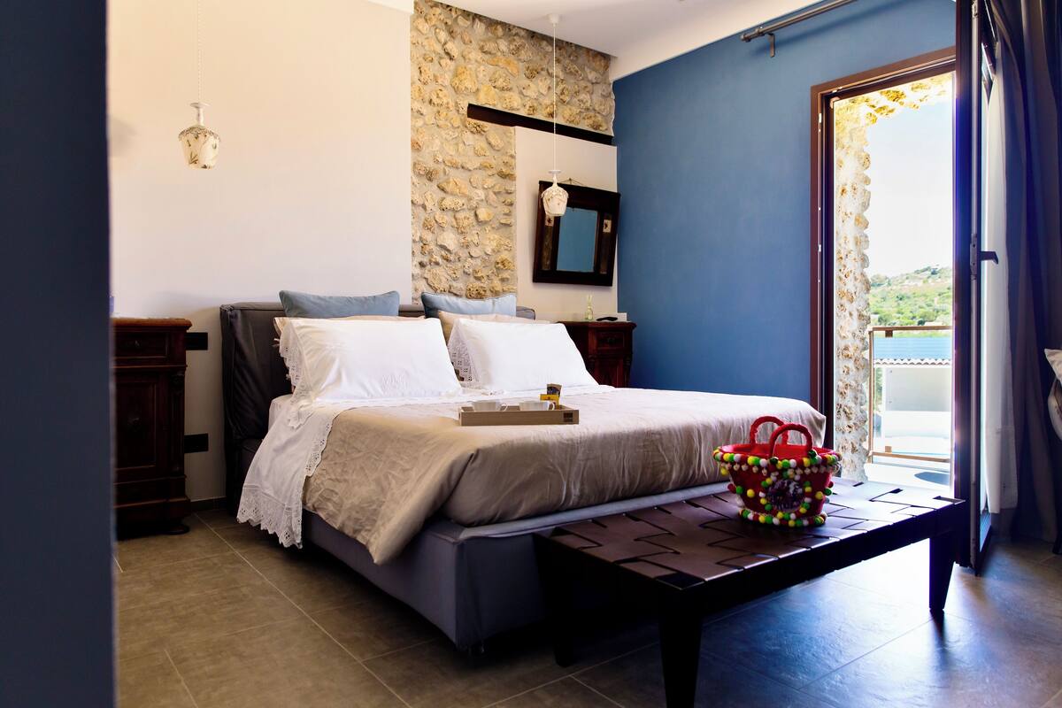 A bedroom with a bed, bedside tables, a stone wall, a mirror, and a view of the outdoors.