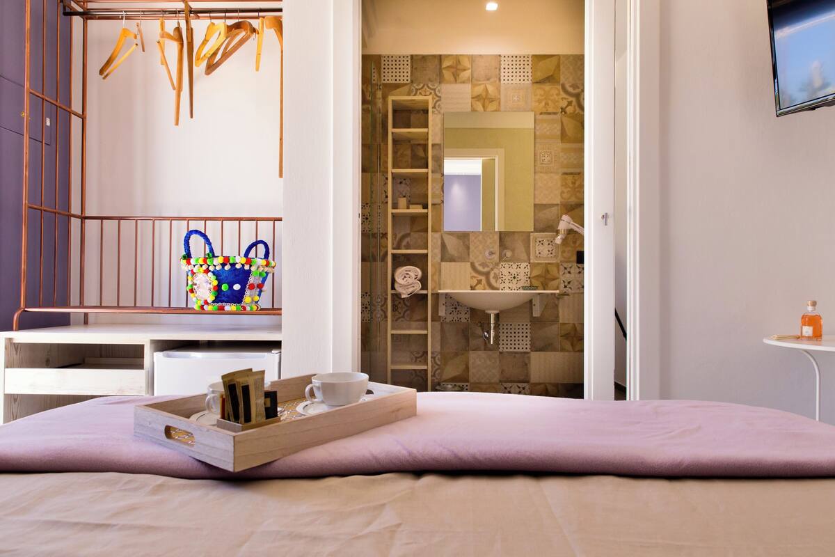 A neatly organized bedroom with a bed, a bedside table with a tray holding a cup and a book, a hanging rack with clothes, and a bathroom visible through an open door.